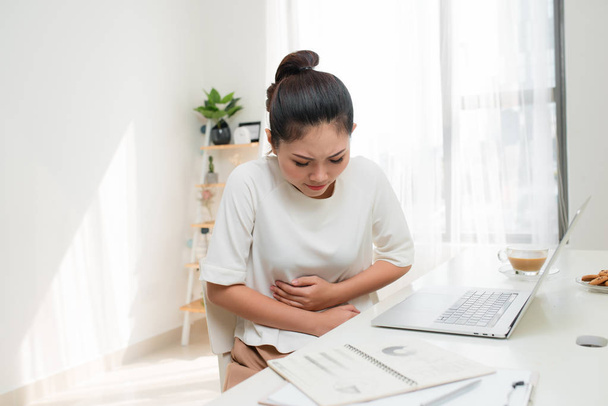 Aasialainen nainen, jolla on vatsakipu tai tunne vatsakipu toimistopöydässä
 - Valokuva, kuva