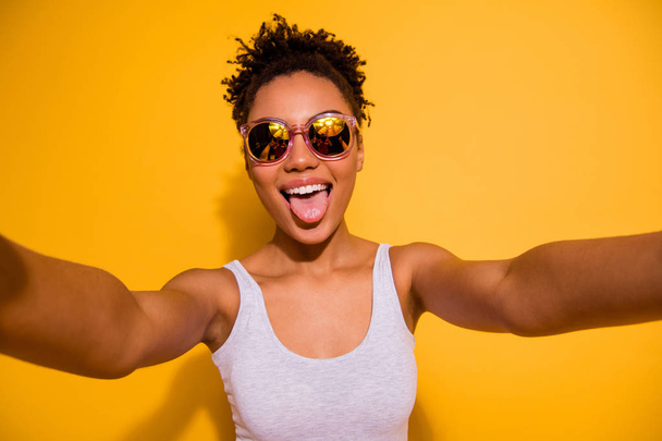 Close up photo beautiful pretty she her dark skin lady summer weekend rest relax make take selfies blogger translation tongue out mouth wear sun specs casual tank-top isolated bright yellow background - 写真・画像