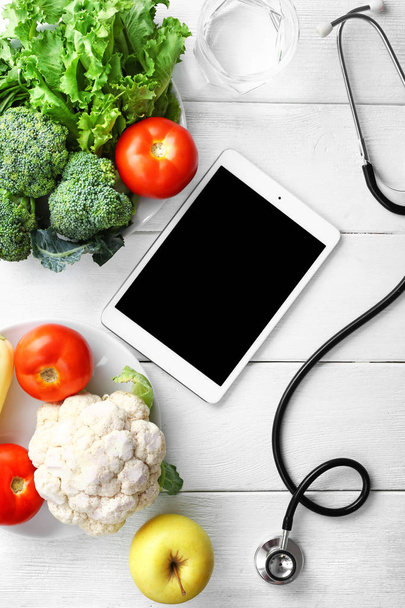 Modern tablet computer with stethoscope and fresh products on white wooden background - Фото, изображение