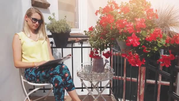 jonge aantrekkelijke vrouw met zonnebril genieten van zon en lezing Magazine op romantisch balkon met bloembakken - Video
