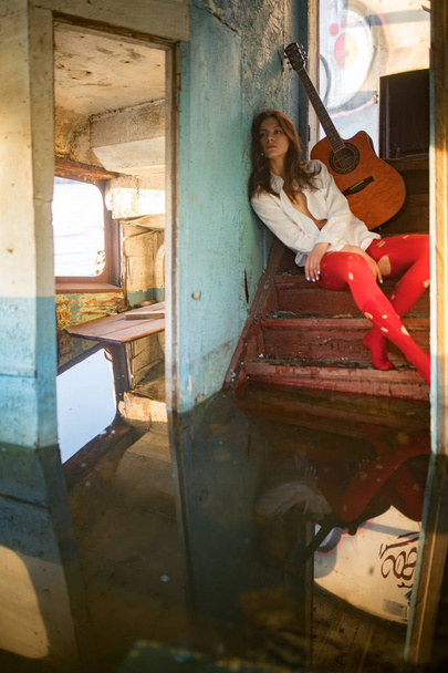 A young sad woman sits with guitar inside of old abandoned ship. - Photo, Image