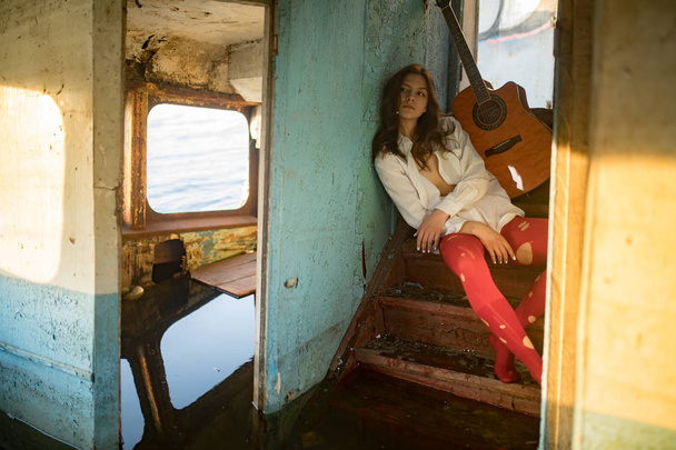 Μια νεαρή θλιμμένη γυναίκα κάθεται με κιθάρα μέσα στο παλιό εγκαταλελειμμένο πλοίο. - Φωτογραφία, εικόνα