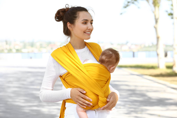 Νεαρή μητέρα με το μωρό της σε σφεντόνα περπατώντας σε εξωτερικούς χώρους - Φωτογραφία, εικόνα