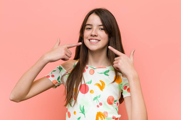 Meisje het dragen van een zomer kleren tegen een rode muur glimlacht, wijzende vingers bij de mond. - Foto, afbeelding