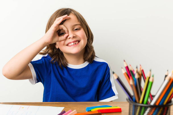 Niño pequeño pintando y haciendo los deberes en su escritorio emocionado mantener el gesto bien en el ojo
. - Foto, Imagen