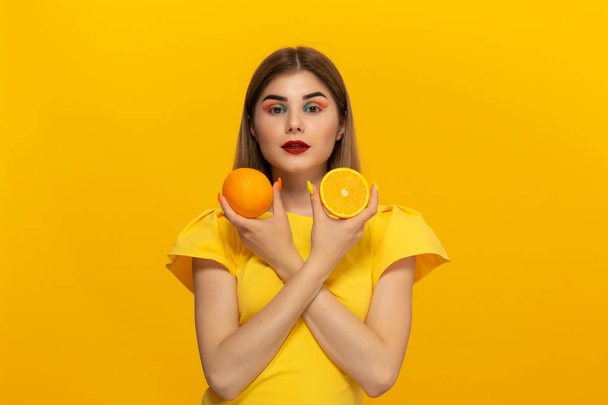 Close-up portrait of a beautiful young girl holding half of oranges close to face isolated over yellow background. - Fotó, kép