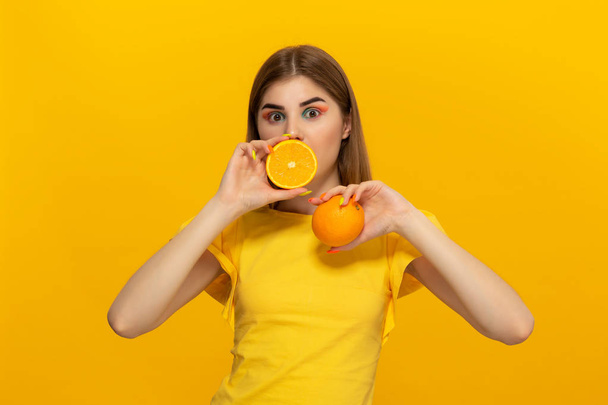 Close-up portrait of a beautiful young girl holding half of oranges close to face isolated over yellow background. - Foto, afbeelding