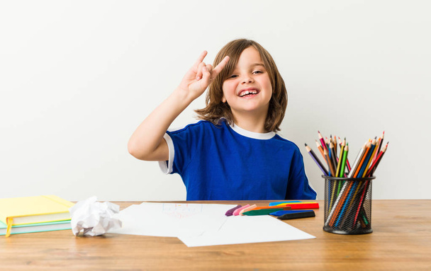 Маленький мальчик рисует и делает домашние работы на своем столе показывая жест рога, как революционная концепция
. - Фото, изображение
