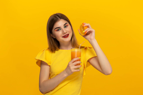 Zbliżenie portret pięknej młodej dziewczyny picie soku pomarańczowego ze szkła gospodarstwa połowę pomarańczowego na białym tle na żółto. - Zdjęcie, obraz