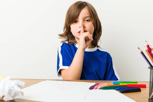 Маленький мальчик рисует и выполняет домашние работы на своем столе, хранит секрет или просит тишины
. - Фото, изображение