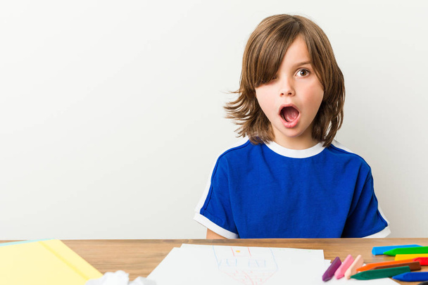 Маленький мальчик рисует и делает домашние работы на своем столе, будучи в шоке от того, что она видела
. - Фото, изображение