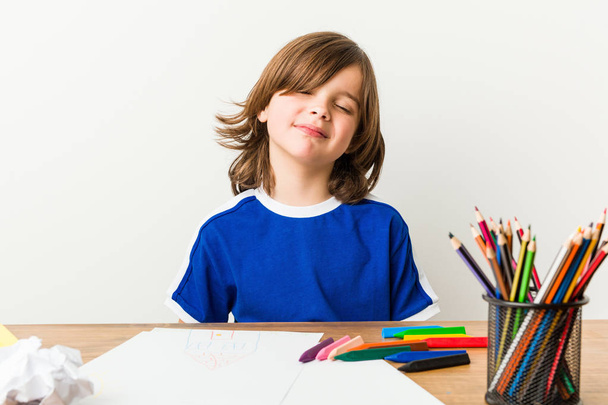 Niño pequeño pintar y hacer los deberes en su escritorio toca la barriga, sonríe suavemente, comer y concepto de satisfacción
. - Foto, Imagen
