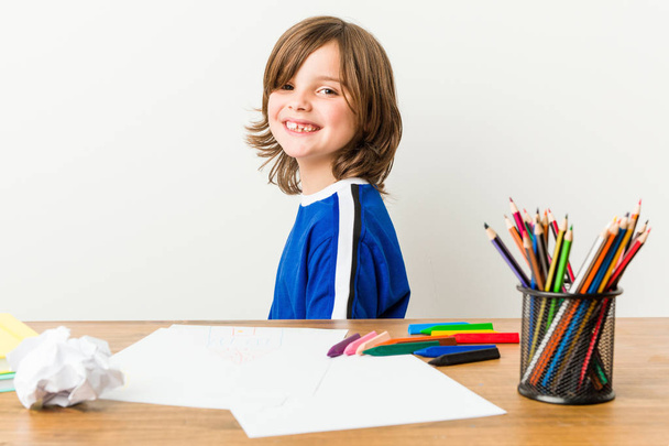 Little boy painting and doing homeworks on his desk looks aside smiling, cheerful and pleasant. - Photo, Image