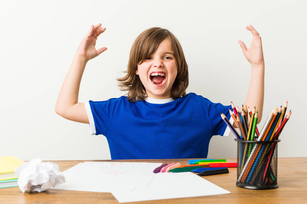 Маленький мальчик рисует и выполняет домашние работы на своем столе, получая приятный сюрприз, взволнован и поднимает руки
. - Фото, изображение