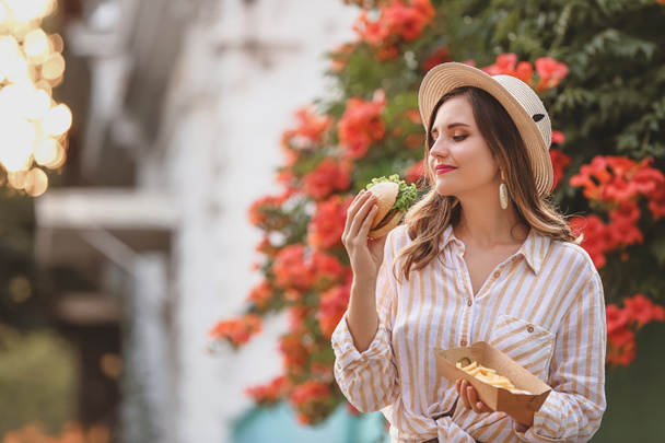 Beautiful young woman eating tasty burger with french fries outdoors - Foto, Bild