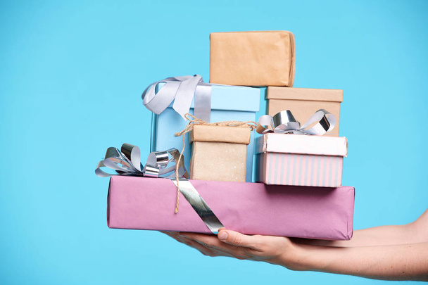 Stapel verpackter und verpackter Geschenke in Schachteln, die isoliert von weiblichen Händen auf blauem Hintergrund gehalten werden - Foto, Bild