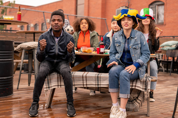 Молодая межкультурная пара футбольных болельщиков и их друзья сидят в кафе на открытом воздухе, пьют пиво с закусками и смотрят матч трансляции
 - Фото, изображение