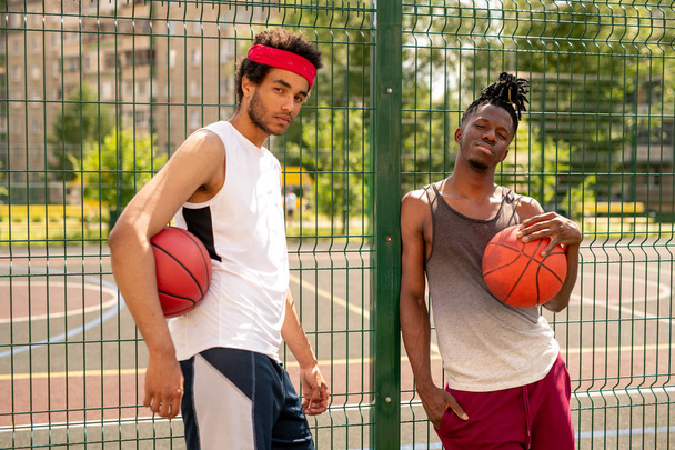 トレーニング後に休息しながら、バスケットボールコートを囲むフェンスのそばに立つスポーツウェアの2人のアクティブな男 - 写真・画像
