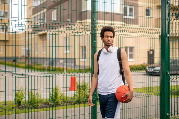 Joven jugador de baloncesto profesional de raza mixta con pelota de pie junto a la valla del patio de recreo en el entorno urbano
 - Foto, Imagen