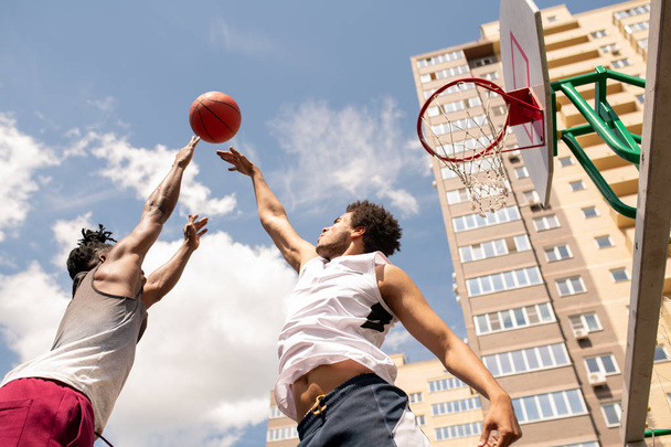 Below view of two intercultural rivals trying to catch ball in motion while training at outdoor court - Fotografie, Obrázek