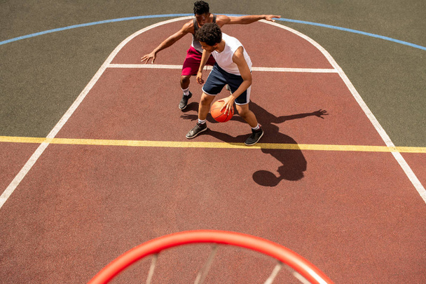 Νέος παίκτης μπάσκετ προσπαθεί να υπερασπιστεί τη μπάλα από την επίθεση του αντιπάλου κατά τη διάρκεια του παιχνιδιού σε υπαίθριο γήπεδο - Φωτογραφία, εικόνα