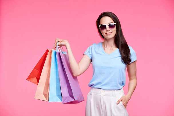 junge stylische Shopaholic in Sonnenbrille und Casualwear prahlt mit ihren Einkäufen, während sie ein paar Papiertüten auf rosa Hintergrund hält - Foto, Bild