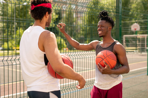 Δύο νέοι πολυπολιτισμικοί παίκτες μπάσκετ που αλληλεπιδρούν μετά το παιχνίδι, ενώ στέκονται από φράχτη - Φωτογραφία, εικόνα