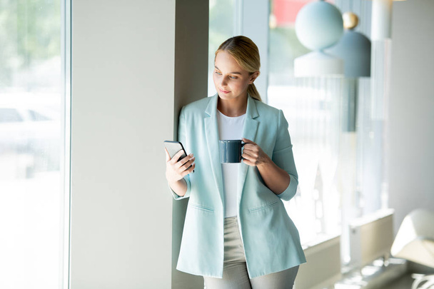 Nuori tyylikäs naispuolinen johtaja lukee ilmoituksen älypuhelimella ja juo kahvia tauolla kahvilassa
 - Valokuva, kuva