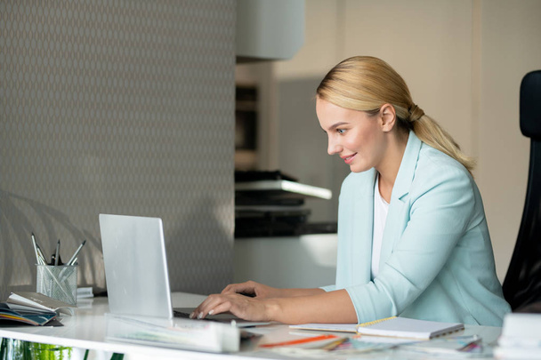 Ευτυχισμένο νεαρό θηλυκό σχεδιαστή κοιτάζοντας την οθόνη του φορητού υπολογιστή, διαβάζοντας ηλεκτρονικά δεδομένα ή εισάγοντας πληροφορίες - Φωτογραφία, εικόνα
