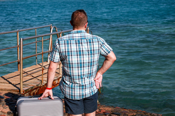 Tourist and traveler is stand on beach of hotel resort with traveled suitcase, enjoys view sea environment, nature landscape, panorama, horizon and rest in sun. Handsome man on vacation in summertime. - Foto, afbeelding