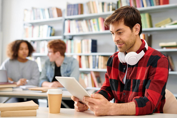 Współczesny uczeń w casualwear oglądanie wideo w Touchpad siedząc przy biurku w klasie - Zdjęcie, obraz