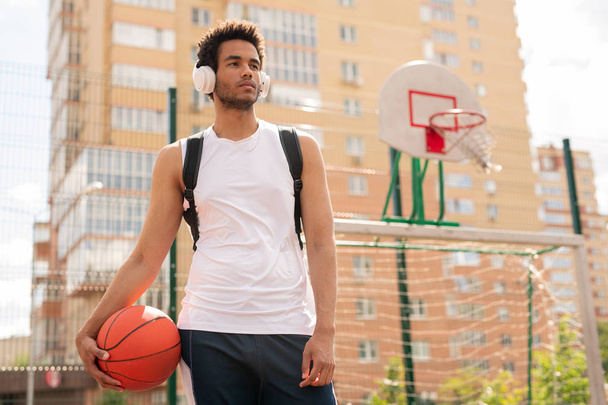 フィールドの遊び場でヘッドフォンで音楽を聴いてバスケットボールをプレイするためのボールを持つスポーティな男 - 写真・画像