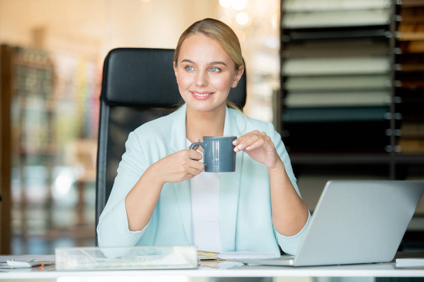 Молода спокійна бізнес-леді з напоєм, сидячи на робочому місці, відпочиваючи перед ноутбуком
 - Фото, зображення