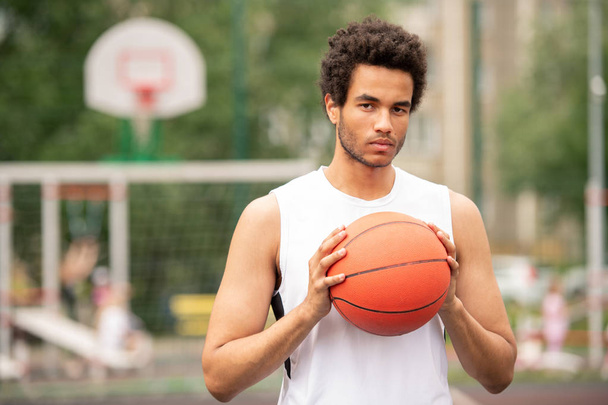 Молодий серйозний мультикультурний спортсмен тримає м'яч для гри в баскетбол, стоячи перед камерою
 - Фото, зображення