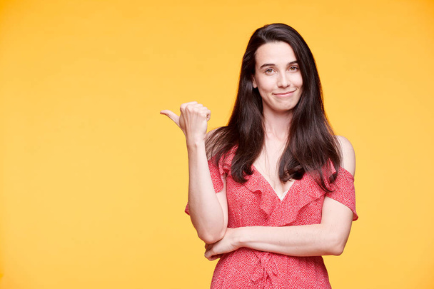 Pretty girl in elegant red dress keeping arms by waist while pointing aside by thumb over yellow background - Foto, Imagem