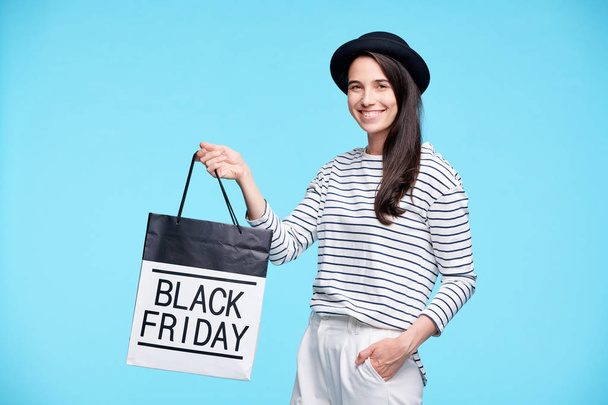 Współczesny wesoły kobieta kupujący w stylowe casualwear pokazując czarny piątek Paperbag patrząc na Ciebie - Zdjęcie, obraz