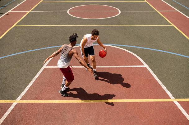 Young basketball player trying to defend ball from rival while carrying it along outdoor court during game - Foto, imagen