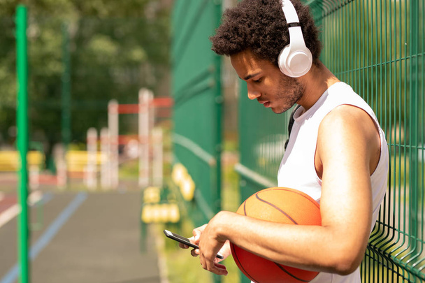 Molada akıllı telefonda kaydırma yaparken çitin yanında duran kulaklıklı genç çağdaş basketbolcu - Fotoğraf, Görsel