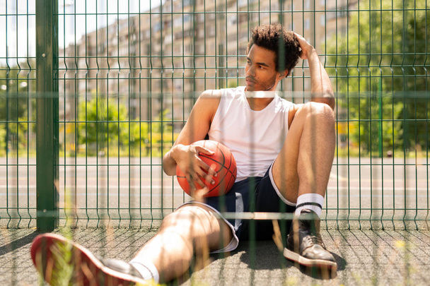 Молодий розслаблений активний чоловік з м'ячем сидить біля паркану баскетбольного майданчика, маючи коротку перерву
 - Фото, зображення