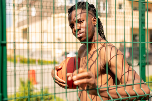 Giovane bel cestista africano che tiene la palla mentre guarda attraverso i bar recinzione che circondano il parco giochi nella giornata di sole
 - Foto, immagini