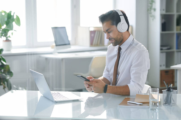 Elegante empleado de oficina en auriculares sentados junto al escritorio mientras busca banda sonora en el teléfono inteligente
 - Foto, imagen