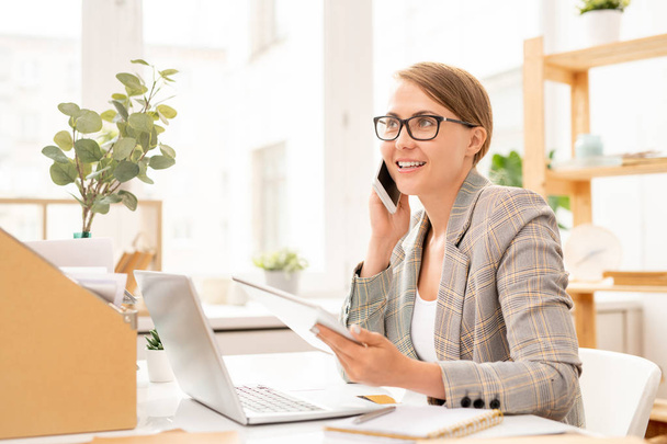 Счастливая деловая женщина разговаривает по смартфону и с помощью планшета перед ноутбуком, сидя за столом в офисе
 - Фото, изображение