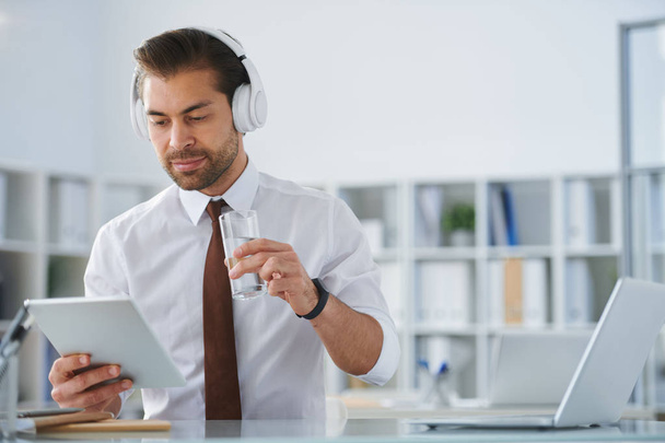 Young serious businessman in headphones looking at tablet screen while having glass of water by workplace - Foto, Imagem