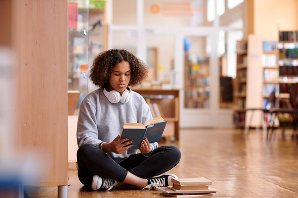 Dość dorywczo uczeń z skrzyżowanymi nogami siedzi na podłodze biblioteki kolegium podczas czytania książki - Zdjęcie, obraz