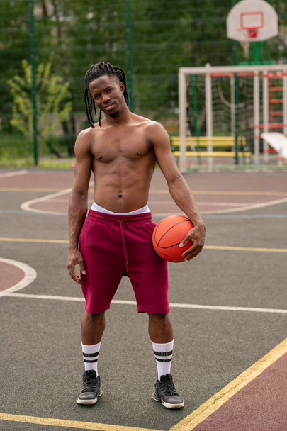 Shirtless African muscular guy with ball standing on basketball court or stadium after training - Φωτογραφία, εικόνα
