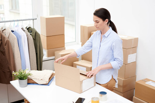 Young office manager in casualwear standing by desk while packing box with online order of client - Photo, Image