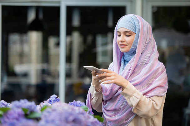Giovane femmina mobile in hijab scattare foto di fiori sulla fotocamera dello smartphone mentre trascorre del tempo all'aperto
 - Foto, immagini