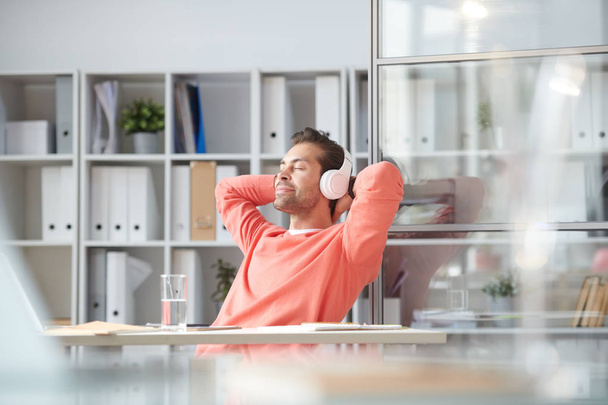 Young relaxed businessman enjoying music in headphones while sitting by desk in office - Zdjęcie, obraz