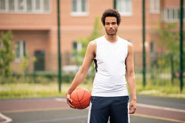 junger gutaussehender Basketballspieler mit Ball, der auf dem Spielfeld vor dem Hintergrund der Architektur steht - Foto, Bild