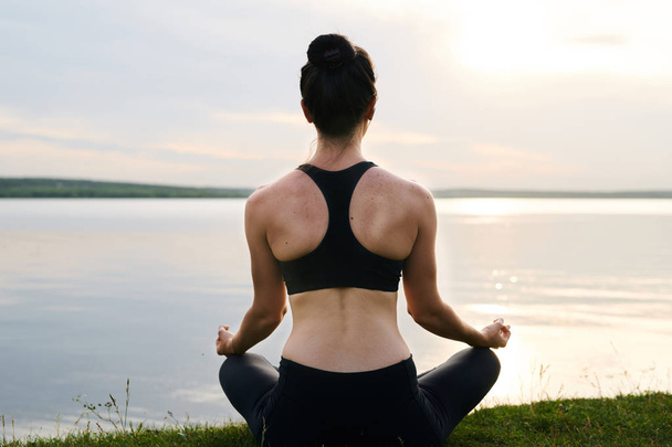 草の上に足を組んで座り、日没時に瞑想する黒いスポーツウェアを着た若い女性の背面図 - 写真・画像